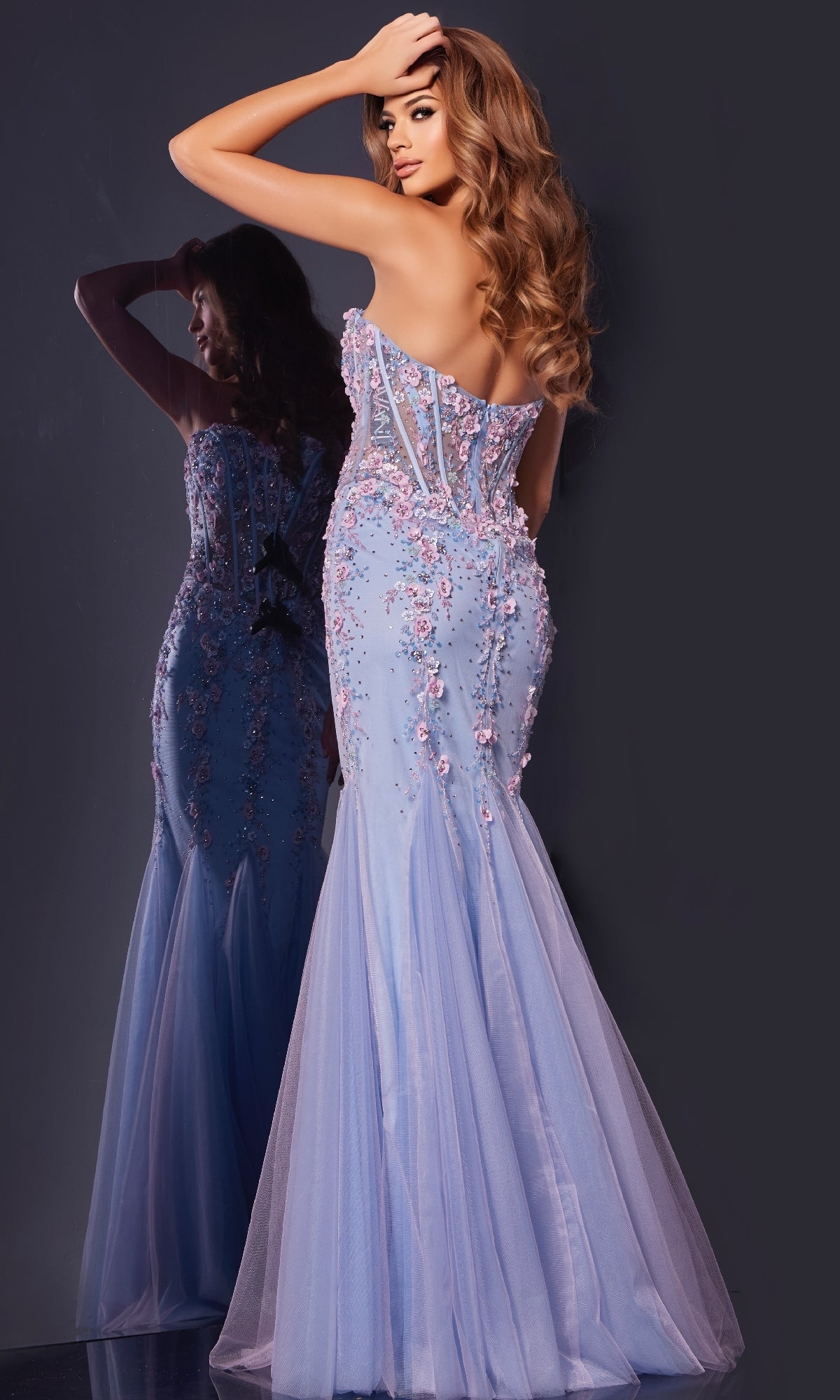 A woman in a Strapless Long Mermaid Prom Dress: Jovani 44211 with floral embellishments stands facing a mirror, showcasing the back of her elegant gown and her flowing wavy hair.